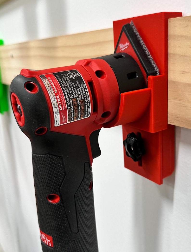 May include: A red and black Detail Sander power tool with a black handle is mounted on a red holder. The tool is attached to a wooden beam. The tool has the text "DETAIL SANDER" and "M12" printed on it.