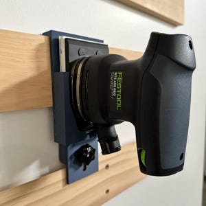 Könnte beinhalten: Eine blaue Wandhalterung für eine Festool RTS 400 REQ Schleifmaschine. Die Schleifmaschine ist schwarz und grün und ist an der Wandhalterung befestigt. Die Wandhalterung hat einen Knopf zum Einstellen des Winkels der Schleifmaschine.