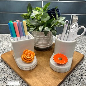 May include: Two white ceramic pen holders with orange flower accents sit on a wooden board. One holds colorful pens, the other holds white scissors and tools. A small potted plant is in the background.