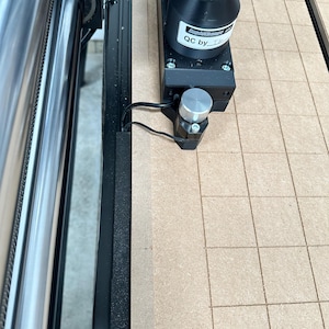 Könnte beinhalten: Nahaufnahme einer CNC-Maschine mit einem schwarzen und silbernen Motor und einem Bürstenkopf. Die Maschine hat eine beige Rasterarbeitsfläche. Der Text "QC by" ist auf dem Motor sichtbar.