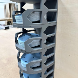 May include: A gray, multi-tiered storage unit filled with stacks of blue sanding discs. The unit has a geometric design with open compartments, providing easy access to the sanding discs. The unit is made of a durable material.