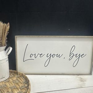 Op de afbeelding: Een rechthoekig houten bord met de tekst "Love you, bye" in zwarte cursieve letters. Het bord heeft een lichtgrijze lijst en een witte achtergrond. Een witte metalen kan en een gevlochten mand staan op de voorgrond.