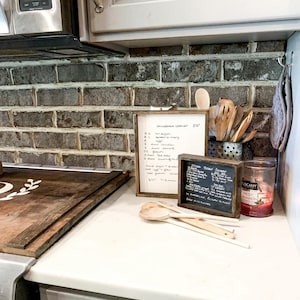 May include: A white countertop with a wooden cutting board, a handwritten recipe list, a small chalkboard sign with a recipe, and wooden spoons. The countertop is in front of a brick backsplash.