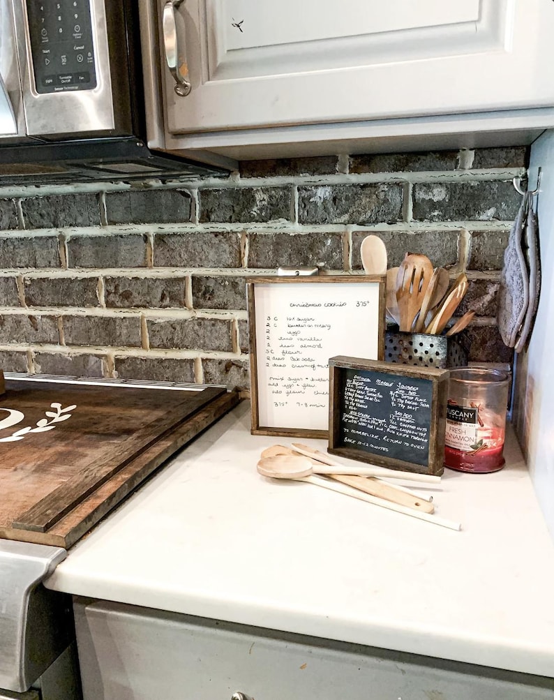 May include: A white countertop with a wooden cutting board, a handwritten recipe list, a small chalkboard sign with a recipe, and wooden spoons. The countertop is in front of a brick backsplash.
