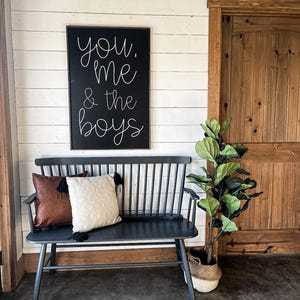 Może przedstawiać: Czarna ławka z brązowym skórzanym poduszką i białą poduszką stoi przed białą ścianą z desek. Czarny znak z białymi literami głosi "you, me & the boys" wisi na ścianie. Roślina w doniczce z zielonymi liśćmi stoi obok ławki.