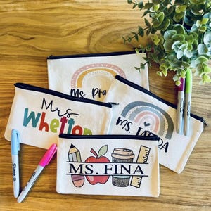 May include: Four white canvas zipper pouches with different designs and teacher names printed on them. The pouches are on a wooden surface with two markers and a plant in the background.