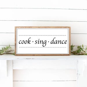 Puede incluir: Un letrero rectangular de madera con las palabras "cook · sing · dance" en escritura negra. El texto está centrado sobre un fondo blanco, enmarcado por un borde de madera marrón. Hay líneas discontinuas encima y debajo del texto. El letrero se muestra en un estante blanco.