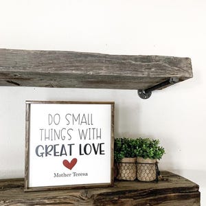 Puede incluir: Un letrero enmarcado con la cita "DO SMALL THINGS WITH GREAT LOVE" en letras negras, un corazón rojo y el nombre "Mother Teresa". El letrero está sobre una estantería de madera rústica con una pequeña planta en maceta.