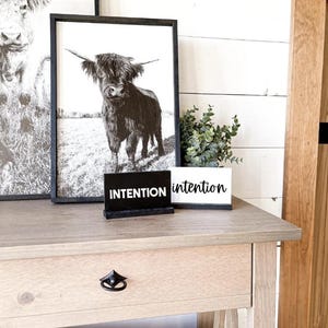 Può includere: Due stampe di mucche delle Highlands incorniciate in nero, una con la parola "intenzione" su un cartello nero e l'altra con la parola "intenzione" su un cartello bianco, appoggiate su un tavolo di legno marrone chiaro con un cassetto.
