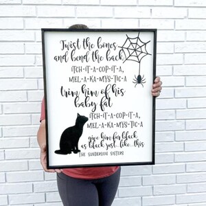May include: A framed sign with a black border and white background. The sign features black text with a Halloween theme, including a spider web, a spider, and a black cat silhouette. The text includes phrases like "Twist the bones" and "The Sanderson Sisters."