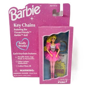 May include: A Barbie key chain featuring the Ocean Friends Barbie doll. The doll is in a pink tutu and has blonde hair. The packaging is pink and features the Barbie logo and the words "Really Works!" and "Basic Fun".
