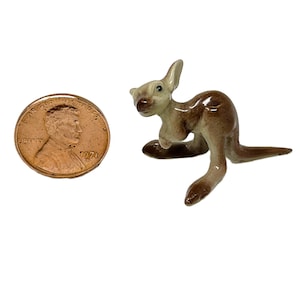 May include: A miniature ceramic kangaroo figurine with a glossy finish, featuring a light cream-coloured head and chest with brown accents on the back, tail, and feet, positioned next to a copper-coloured United States one-cent coin for scale.