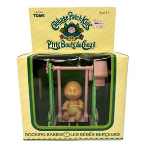 May include: A Cabbage Patch Kids Rocking Babies toy in its original yellow packaging. The set includes a small, bald doll wearing a nappy, seated on a pink swing set with green side supports. The box features the text Cabbage Patch Kids, P'tits Bouts de Choux, and Rocking Babies Les Bébés Berceurs.