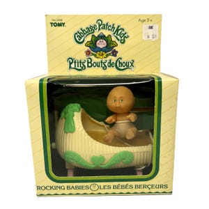 May include: A Cabbage Patch Kids Rocking Babies toy set. The box features the Cabbage Patch Kids logo and the text "Ptits Bouts de Choux". Inside, a small doll sits in a cream-coloured cradle with green accents. The box is yellow with green trim.