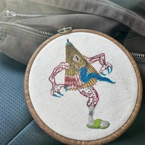 Può includere: Arte di ricamo a cerchio con un personaggio dei cartoni animati con un corpo giallo, marrone e rosa, una sciarpa blu e una pantofola verde. Il personaggio ha un occhio grande e una lingua lunga.