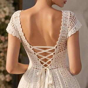 May include: A white crochet dress featuring short sleeves and a deep, open back design. The back is secured with a crisscross ribbon lacing system that ties into a bow at the waist. The garment is crafted with an intricate, openwork knit pattern.
