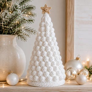 May include: A white crocheted Christmas tree decoration with a textured bobble stitch pattern and a small, light brown crocheted star topper. The conical tree stands upright, featuring a flat, circular base and a dense, uniform surface texture.