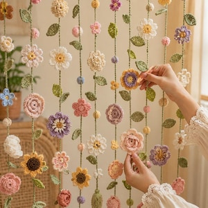 May include: A decorative hanging garland featuring multiple vertical strands of crocheted flowers and leaves in various colors including pink, yellow, white, purple, and green. The floral elements are attached to thin green yarn strings.