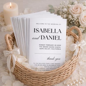 May include: A woven basket holds wedding programs with the names Isabella and Daniel. The programs are white with black text, including the wedding date and location. The basket is decorated with white ribbons and surrounded by flowers and a lit candle.