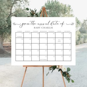 May include: A white sign on a wooden easel with the text "guess the arrival date of BABY CHARLIE." The sign is a calendar grid for August and September, with dates and days of the week. Decorative hearts are at the top.