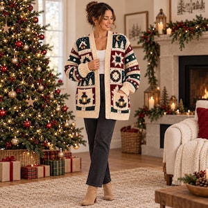 May include: A long-sleeved, open-front crochet cardigan featuring a festive patchwork design with motifs of pine trees, houses, stars, and lanterns in shades of cream, dark green, and deep red, worn over a white top and dark gray denim jeans.