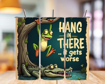 May include: A cylindrical tumbler featuring a cartoon illustration of a green frog hanging from a tree branch above a crocodile in water. The text HANG IN THERE .. it gets worse is printed in yellow. The design includes a metal straw at the top.