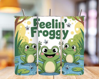 May include: A white tumbler with a cartoon frog design. The tumbler features three green frogs in a pond with the words "Feelin' Froggy" in green. The background includes trees, grass, and flying insects. A silver straw is visible.