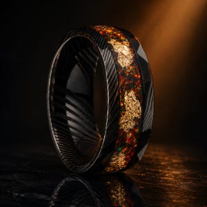 Puede incluir: Un anillo negro con una banda metálica oscura y texturizada, que presenta una incrustación central de fragmentos de ópalo rojo y verde iridiscente, intercalados con copos de pan de oro irregulares y brillantes sobre un fondo oscuro.