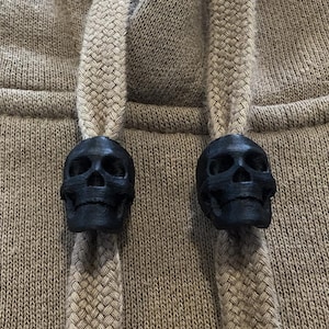 Puede incluir: Dos botones de ajuste de capucha en forma de calavera negra. Los botones están unidos a cordones de ajuste de color beige.