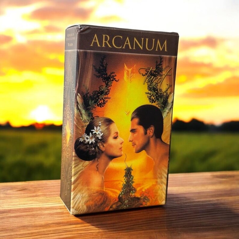 May include: A tarot card box with the word "ARCANUM" at the top. The box features an illustration of a man and woman facing each other, with a golden background. The word "Tarot" is also visible. The box is sitting on a wooden surface.