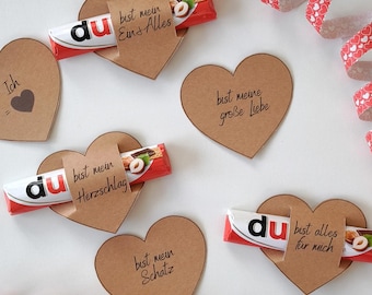 May include: A collection of brown heart-shaped cards with German text, paired with Kinder Bueno chocolate bars. Decorative red and white heart-patterned ribbons are scattered around the cards. The cards have phrases like "bist mein Schatz" and "bist meine große Liebe".