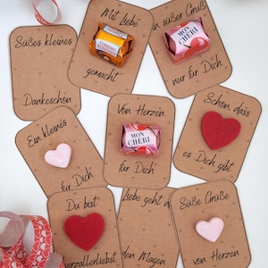 Puede incluir: Una colección de etiquetas de regalo marrones con frases en alemán y adornos en forma de corazón. Algunas etiquetas presentan caramelos como Ferrero Rocher y Mon Cheri. También hay una cinta roja y blanca con patrones de corazón.