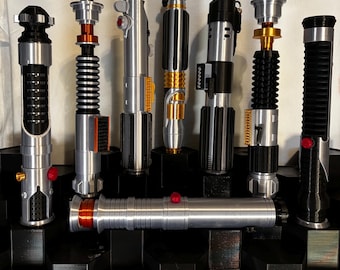 May include: A collection of seven lightsaber hilts displayed on a black tiered stand. The hilts feature various metallic finishes including silver, black, and gold, with cylindrical designs, ribbed grips, and small red or gold buttons and accents.