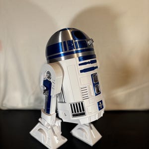 May include: A white and blue robotic figure with a domed silver head. The cylindrical body features moulded panel details, including rectangular vents, blue accents, and two side legs with flat feet. The design mimics a classic astromech droid.