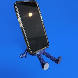 Può includere: Un supporto per telefono blu navy a forma di persona, con braccia e gambe. Sostiene uno smartphone nero con una custodia trasparente. Lo sfondo è blu unito.