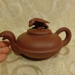 May include: A terracotta teapot with a bird-shaped lid and a snake-shaped spout. The teapot is a reddish-brown color and has a smooth, matte finish.
