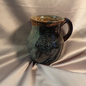 May include: A handmade ceramic mug with a dark brown handle and rim. The mug's body features a speckled pattern in shades of blue, black, and green. The interior and rim have a brown and orange glaze. This unique mug is perfect for coffee or tea.