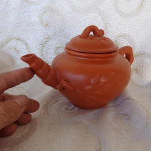 May include: A small, terracotta teapot with a lid and a leaf design. The teapot is about the size of a person's hand.