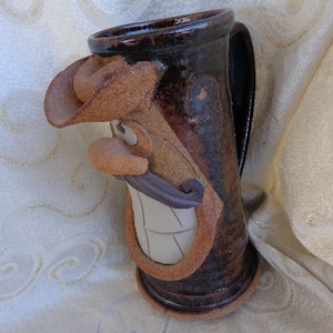 May include: A brown ceramic mug with a whimsical face, featuring a wide smile, a mustache, and a cowboy hat. The mug has a handle and a dark brown glaze.