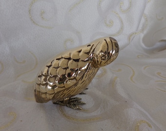 May include: A gold-colored metal bird figurine with a detailed, textured surface. The bird is perched on its feet and has its head tilted down.