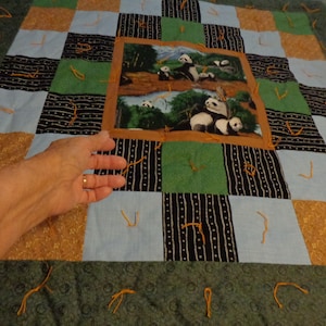 May include: A patchwork quilt with a central panel featuring a colorful image of pandas in a forest setting. The quilt is made of various fabrics in shades of green, brown, black, and blue. The quilt is tied with orange yarn.