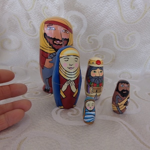 May include: A set of six hand-painted wooden nesting dolls depicting the Nativity scene. The dolls are painted in bright colors and feature detailed faces and clothing. The largest doll is of Joseph, followed by Mary, the three wise men, and the baby Jesus.