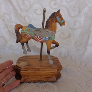 May include: A brown carousel horse figurine with a blue saddle and an American flag design. The horse is on a wooden base with a gold pole.