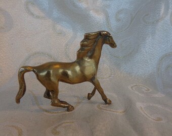 May include: A small, gold-colored metal horse figurine. The horse is standing on all four legs with its head turned to the side. The horse has a flowing mane and tail.