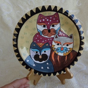 May include: A ceramic plate with a black and gold rim features three cats with colorful, whimsical designs. The cats are stacked on top of each other, with the top cat being a pink and white cat with black spots, the middle cat being a blue and black cat, and the bottom cat being a brown and gray cat with orange accents.