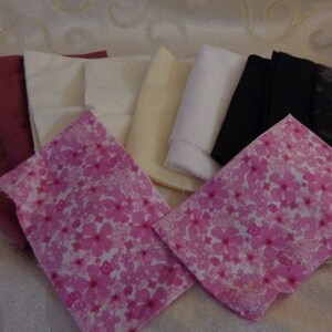 May include: A collection of folded fabric squares in various colors and patterns. The assortment includes solid colors like burgundy, cream, white, and black, alongside a pink floral print. The fabrics are neatly arranged, showcasing their textures and hues.