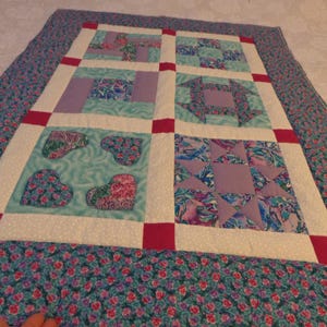 May include: Handmade quilt with a patchwork design. The quilt features squares with heart, geometric, and star patterns in shades of blue, purple, and pink. The border is a floral print in blue and pink, with a white dotted background.