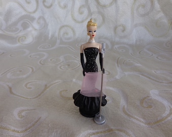 May include: A black and white Barbie doll ornament wearing a black sequined dress and a pink shawl, holding a microphone on a stand.