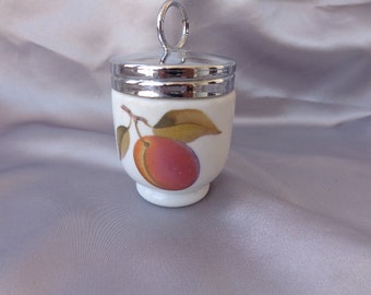 May include: A white ceramic egg cup with a silver lid and a painted image of a peach with green leaves.