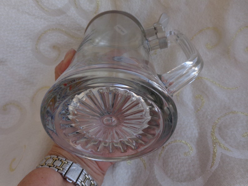 May include: A clear glass beer stein with a pewter lid and a handle. The bottom of the stein has a decorative, starburst pattern.